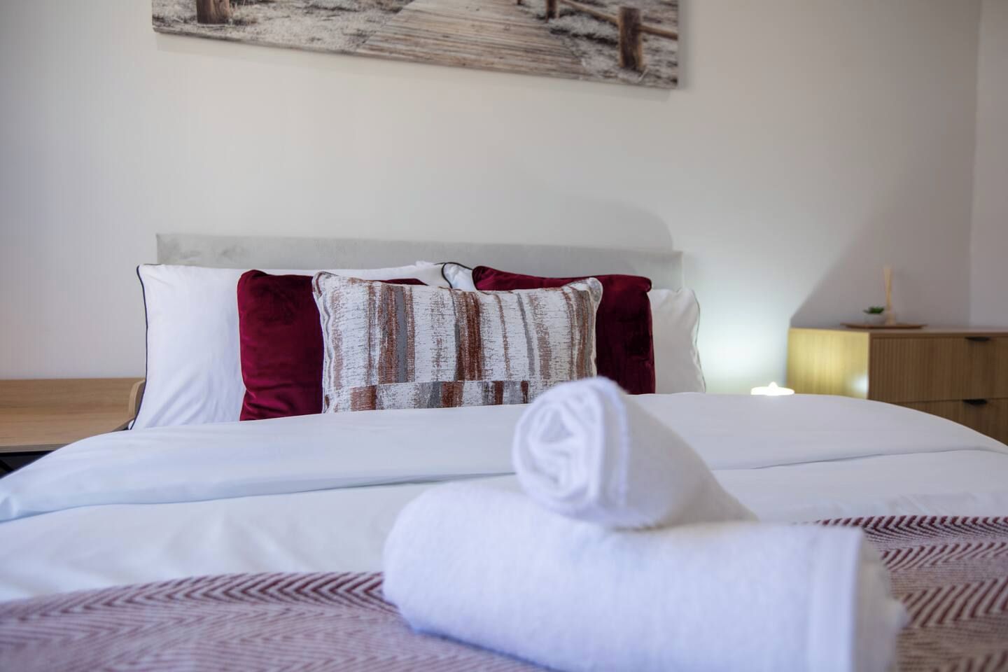 A neatly made bed with white linens and a rolled towel, a wooden nightstand with a lamp, and a wall with a rustic painting.