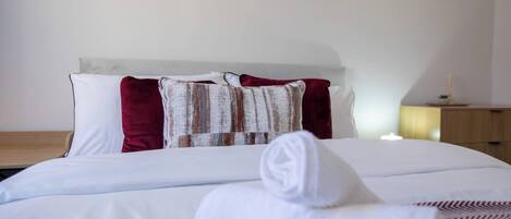 A neatly made bed with white linens and a rolled towel, a wooden nightstand with a lamp, and a wall with a rustic painting.