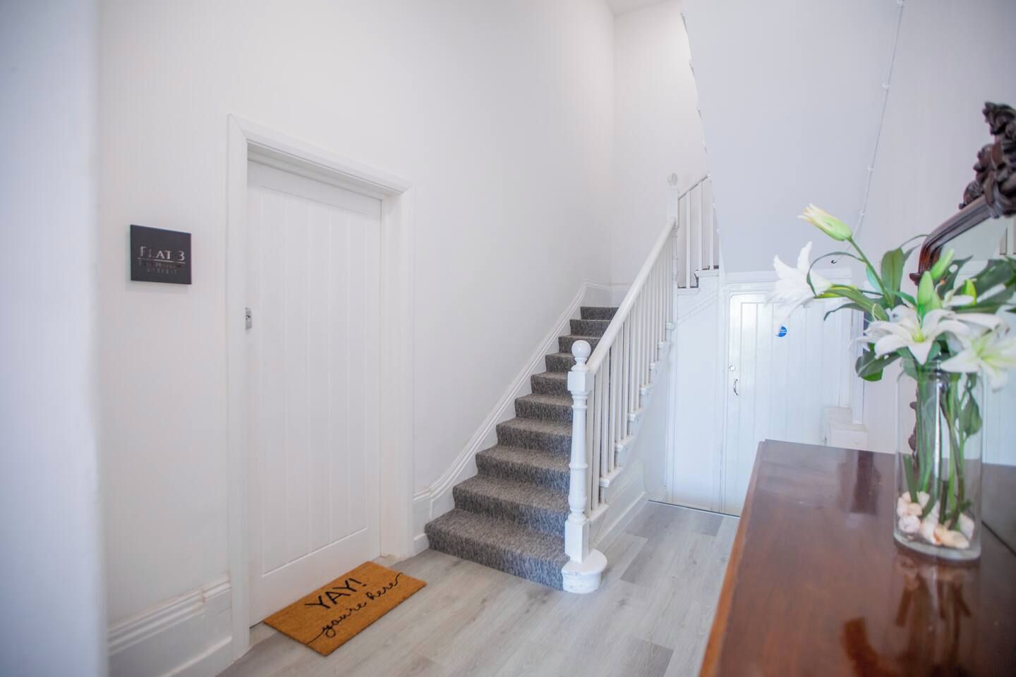 Un escalier avec une rampe blanche, un paillasson sur lequel on peut lire les mots « HOURRA » et « vous êtes là », et un vase rempli de fleurs posé sur une table.