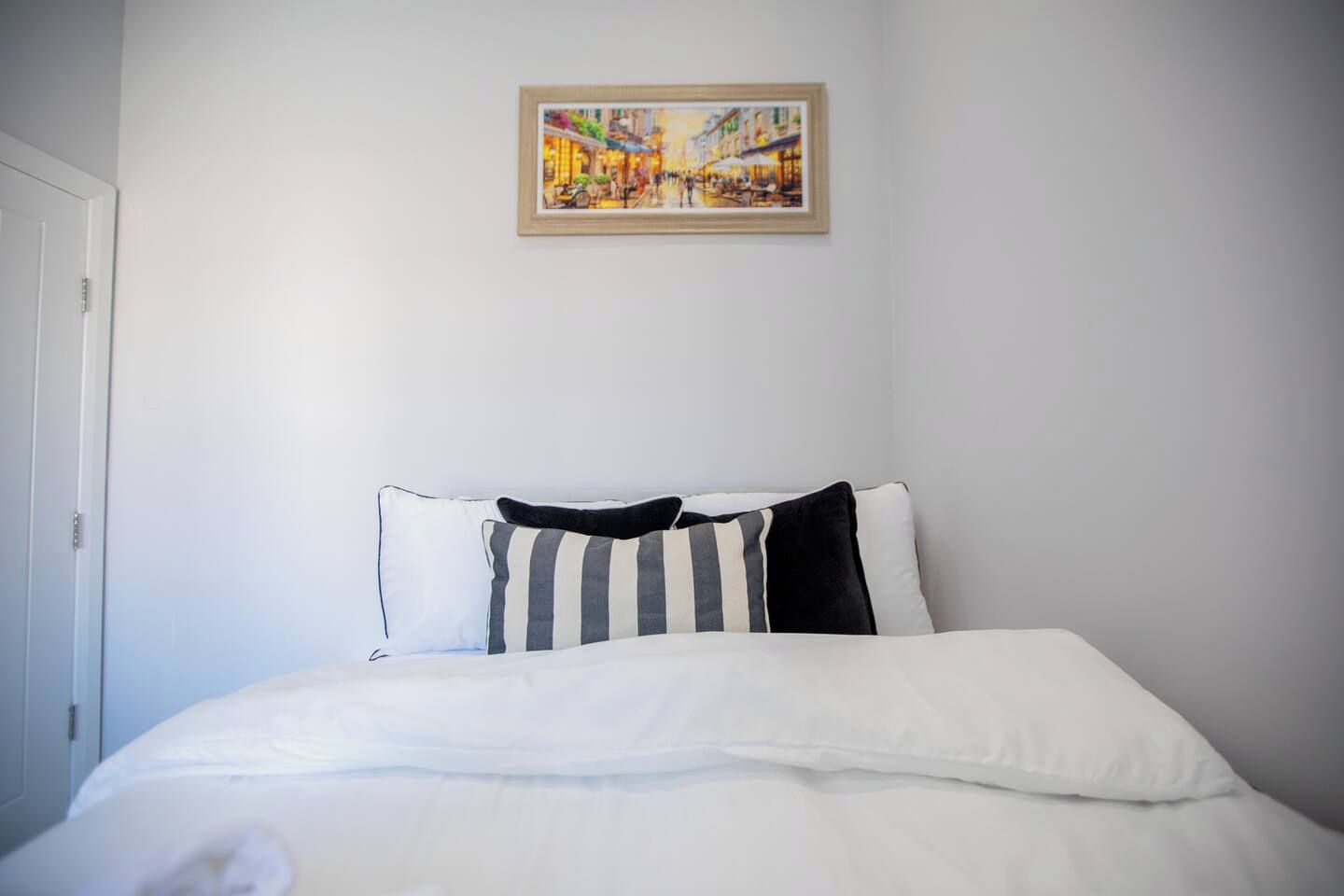 Una habitación de hotel con cama, ropa de cama blanca, almohada a rayas y un cuadro en la pared.
