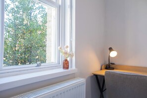 Ein Zimmer mit Fenster, Heizkörper, Schreibtisch mit Lampe und einer Blumenvase auf der Fensterbank.