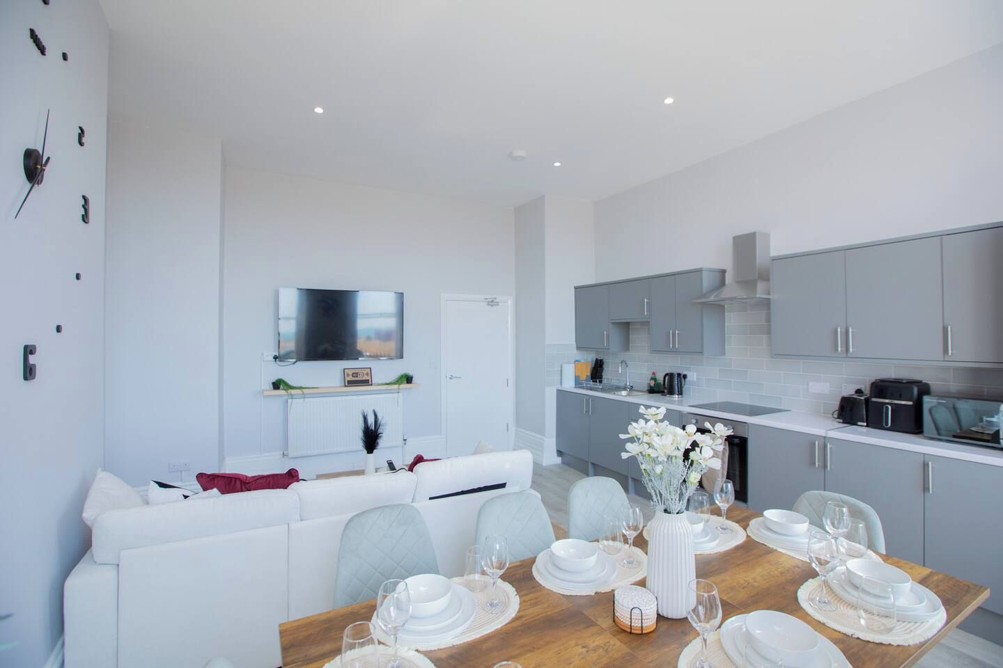 A modern kitchen with grey cabinets, a dining area with a wooden table set for four, and a living area with a white sofa.