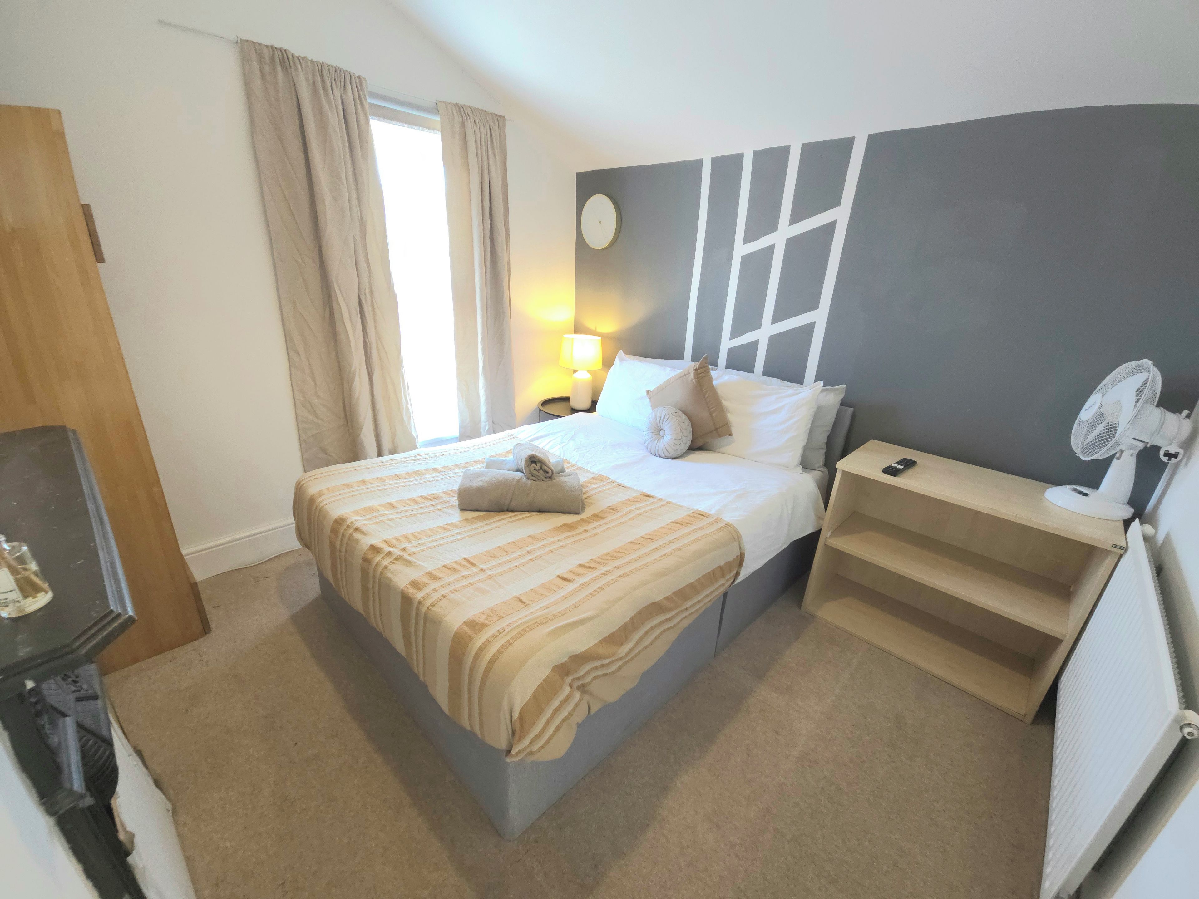 A hotel room with a bed, bedside table, lamp, and a standing fan.