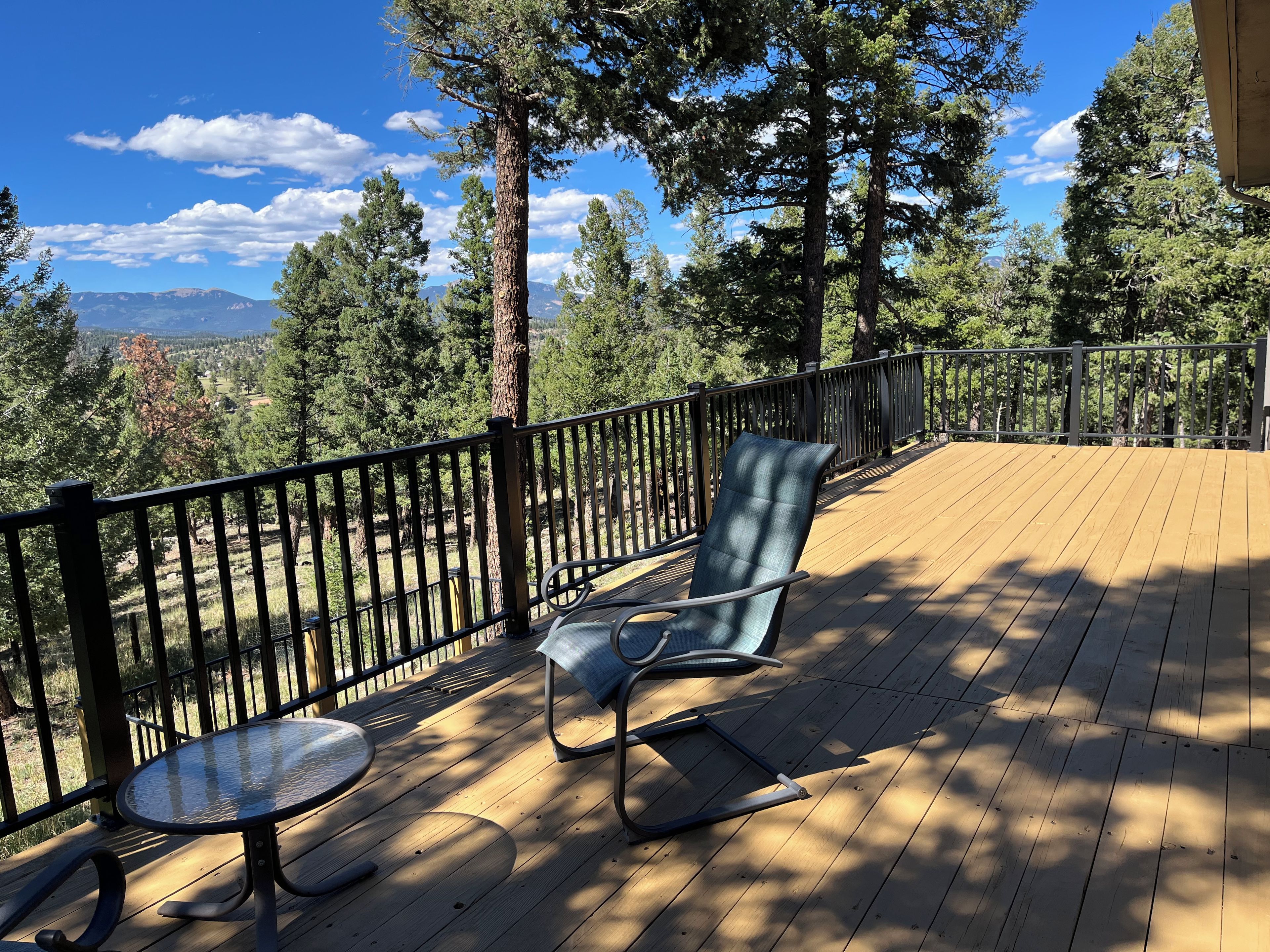 Una terraza de madera con dos sillas y una mesa, con vistas a una zona boscosa y montañas a lo lejos.