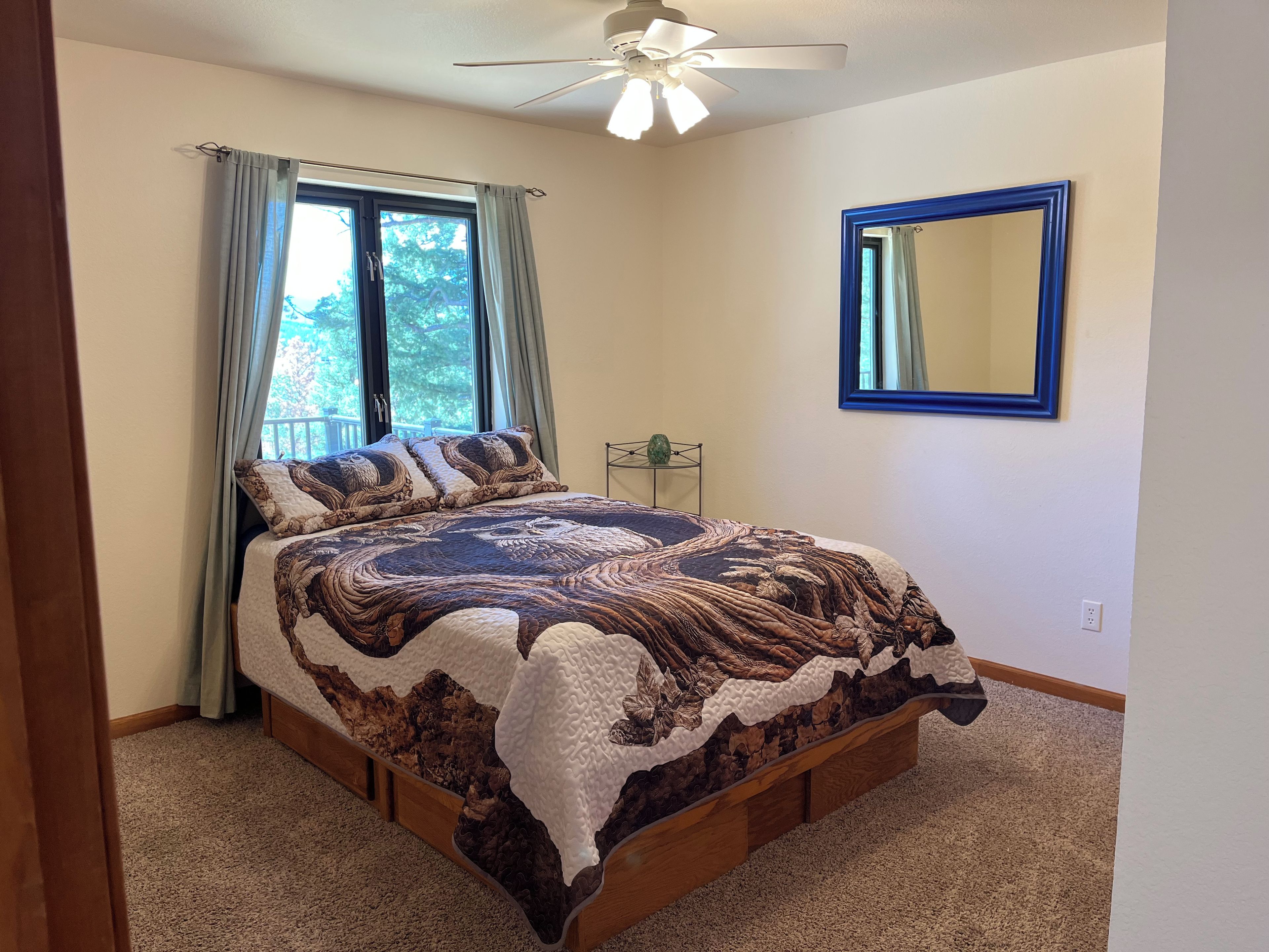 A bedroom with a bed, a ceiling fan, a window with curtains, a mirror, and a small table with a vase.