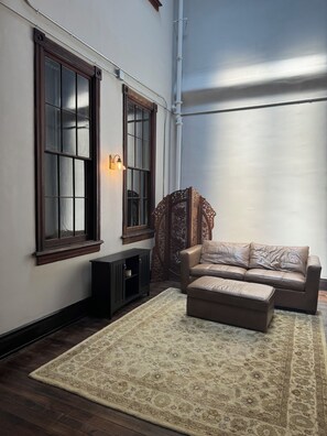 A room with a large window, a sofa, a matching ottoman, a dark wooden cabinet, and a patterned rug.