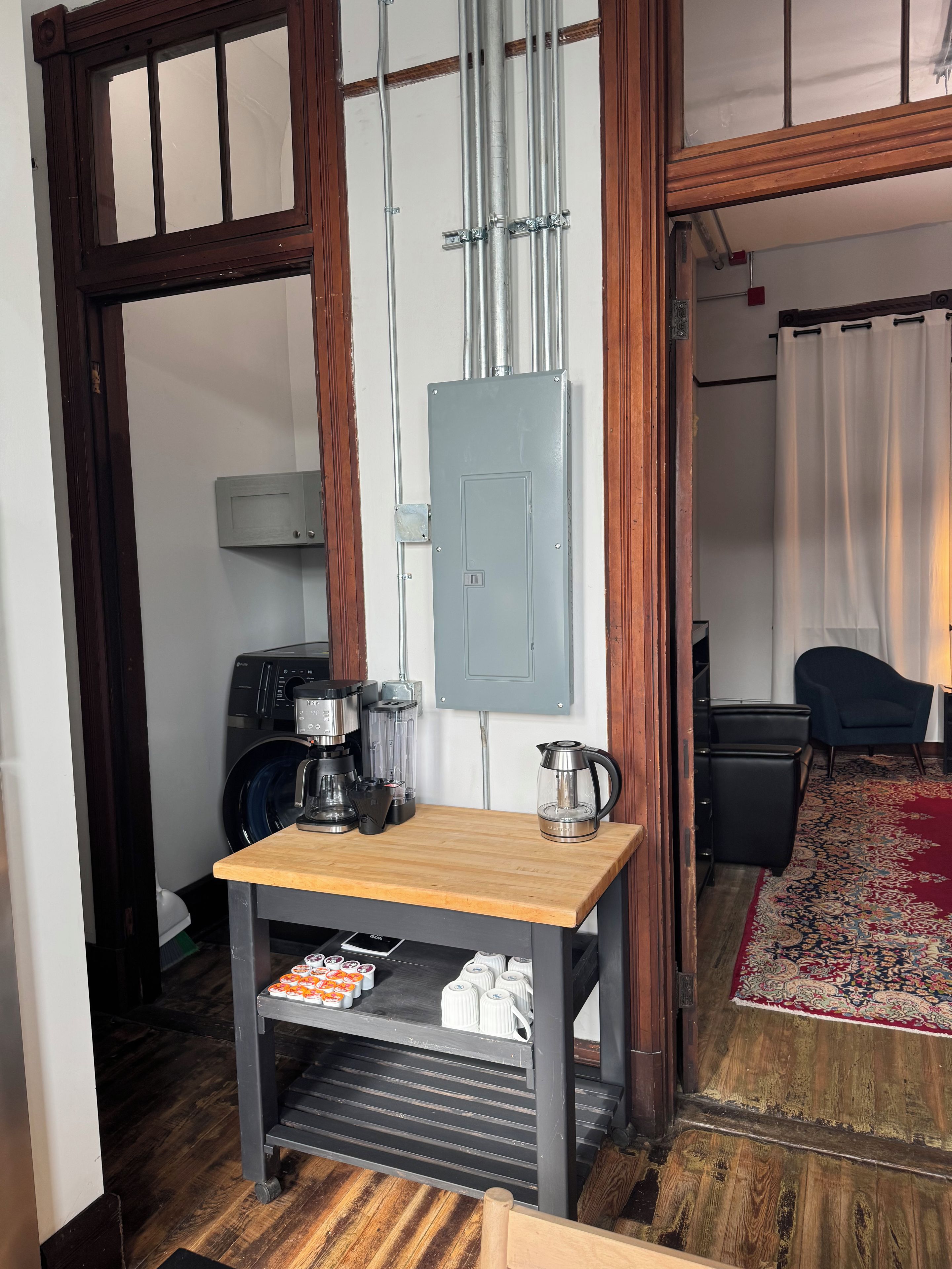 A compact room with a wooden table, a washing machine, a coffee maker, and a power outlet box.