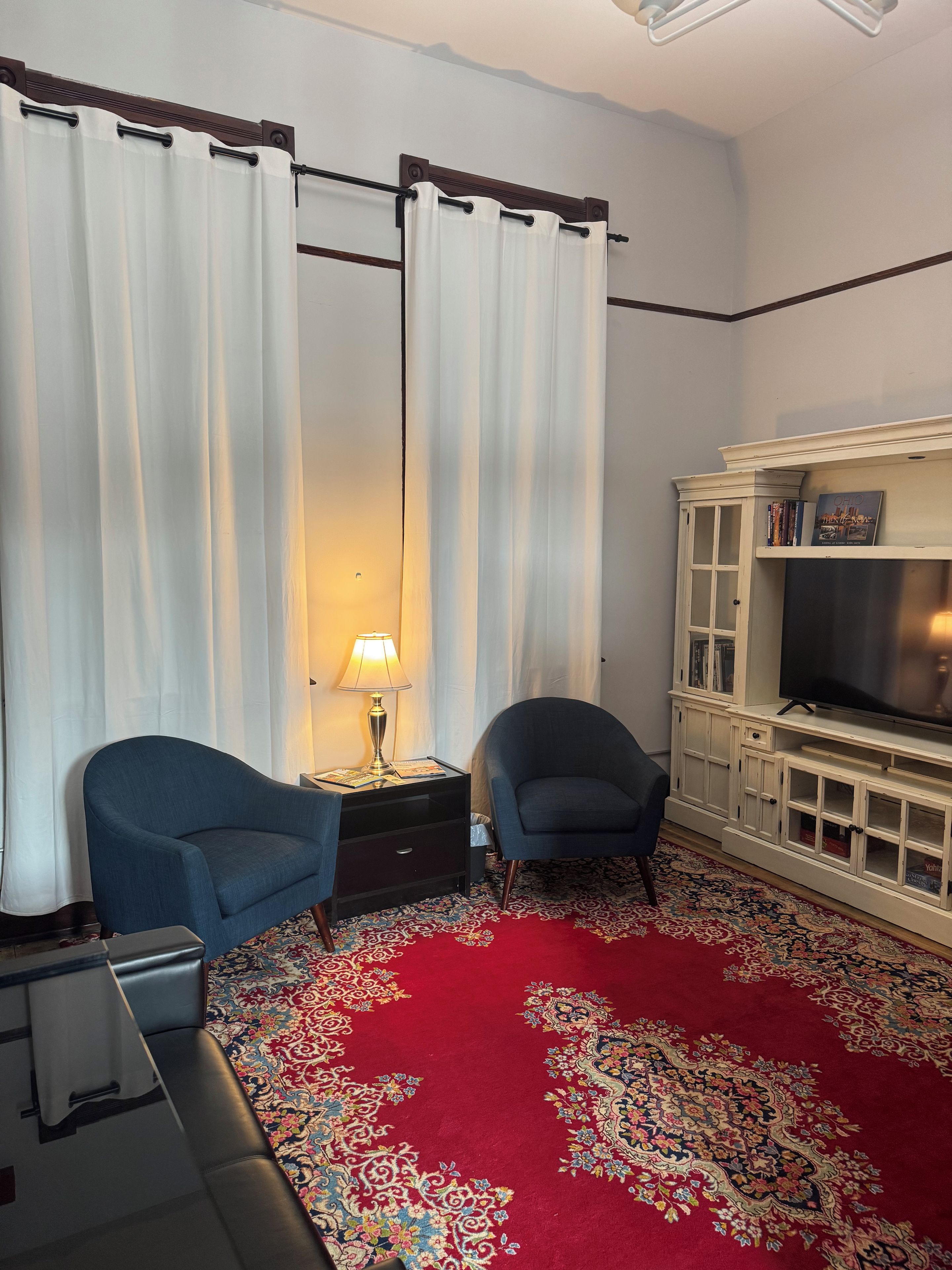 Ein Zimmer mit einem roten Teppich, zwei Sesseln, einem Fernseher auf einer weißen Kommode und einer Lampe auf einem Beistelltisch.