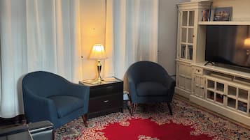 A room with a red patterned carpet, two armchairs, a television on a white cabinet, and a lamp on a side table.