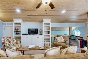 A living room with a wooden ceiling, a flat-screen TV, white shelving units, and a window with a view.