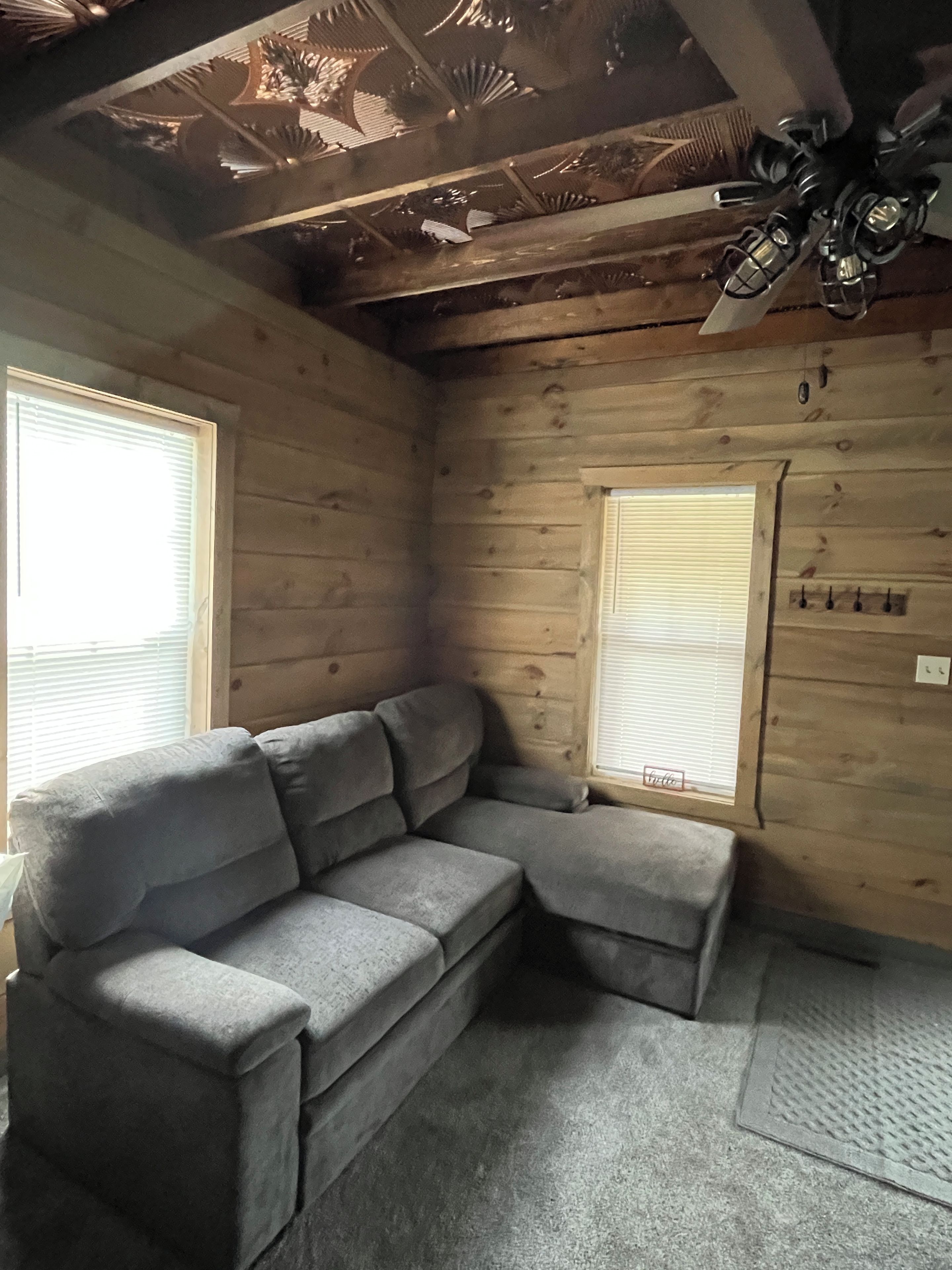 A room with a gray sectional sofa, wooden walls, and a ceiling fan.