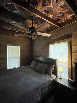 A bedroom with a bed, a nightstand, a lamp, a ceiling fan, and a window with blinds. - Cozy Log Cabin Retreat (Macon)