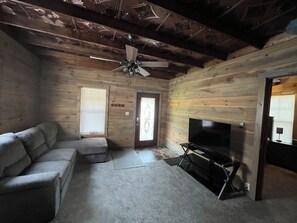 A log cabin interior with a sofa, a fireplace, a ceiling fan, and wooden walls. - Cozy Log Cabin Retreat (Macon)