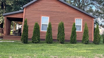 A log cabin with a porch, two windows, and six coniferous trees in front.