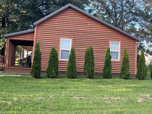 A log cabin with a porch, two windows, and six coniferous trees in front.