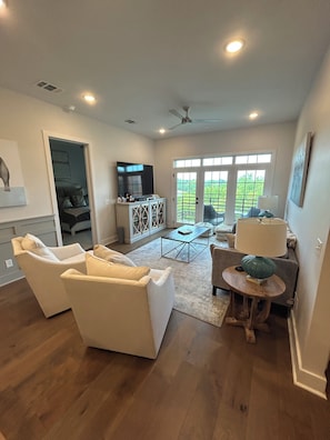 A spacious living room with a flat-screen TV, a ceiling fan, and a large window with a view of greenery. - Grand Junction Football Condo! Walkable to Stadium and golf cart service. (Starkville)
