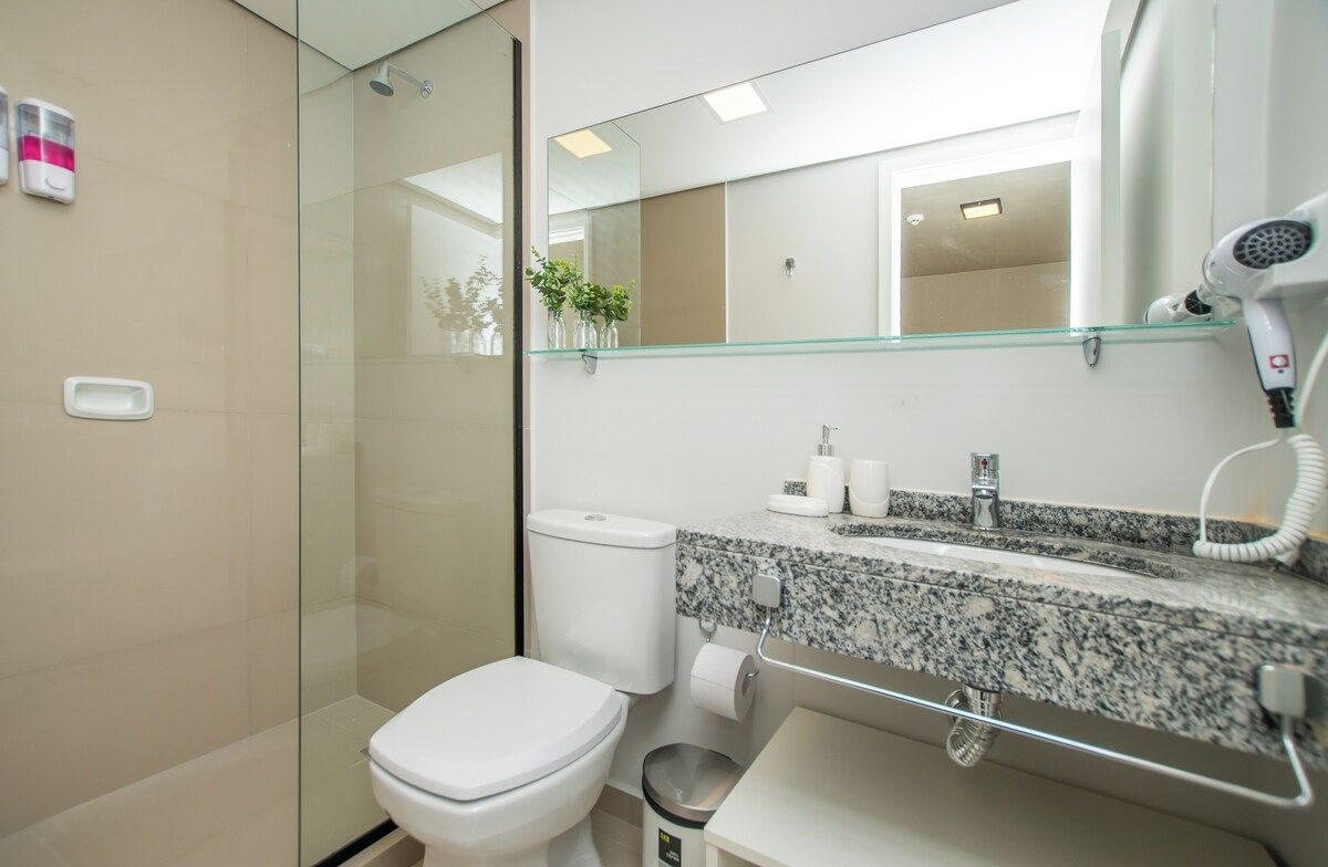 Een moderne badkamer met een douchecabine van glas, een wit toilet, een granieten aanrechtblad, een haardroger en een spiegel.