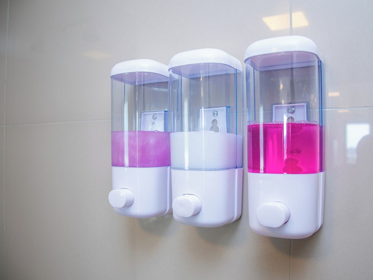 Três dispensadores de sabonete fixados na parede, com partes transparentes e líquidos coloridos.