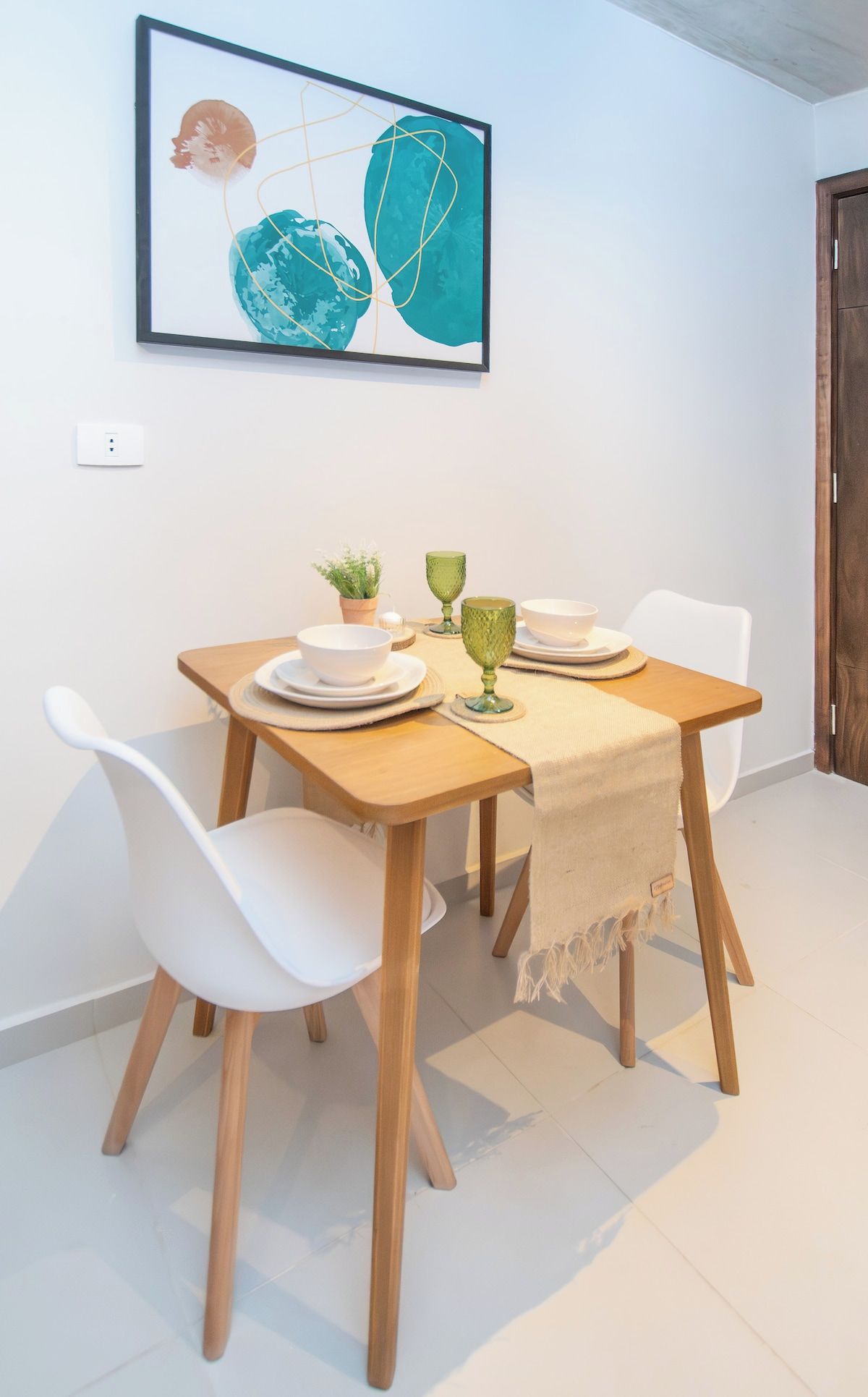 A dining area with a table set for two, white chairs, a small potted plant, and a framed artwork above.