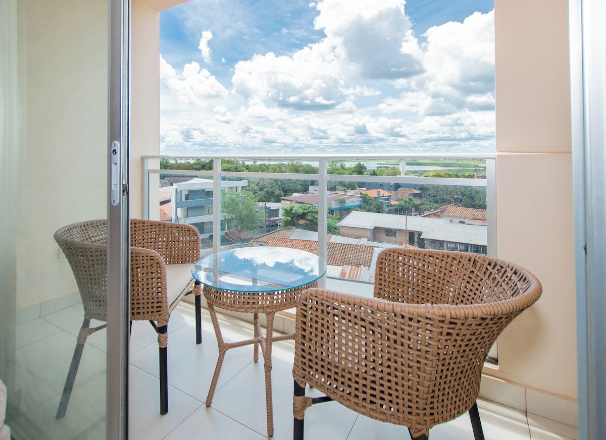 A balcony with wicker furniture and a glass table, overlooking a residential area with houses and a clear sky.