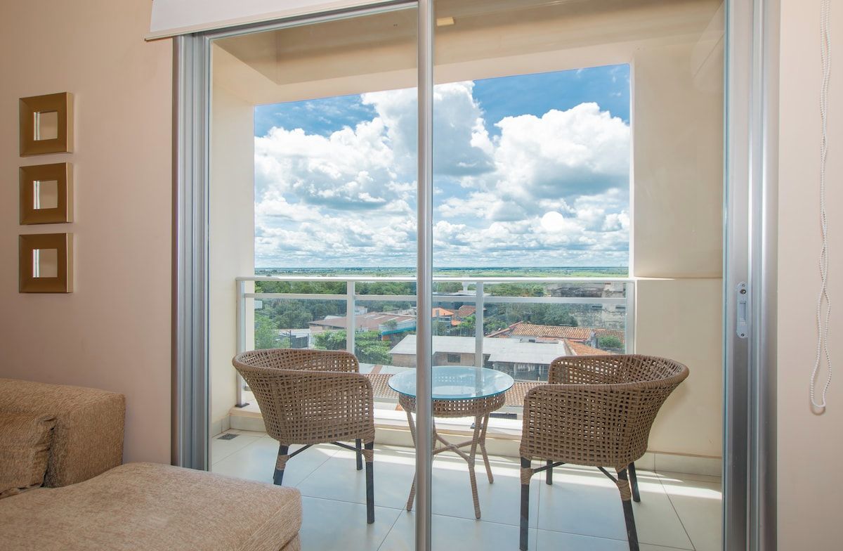 A balcony with wicker chairs and a glass table, overlooking a cityscape and a cloudy sky.