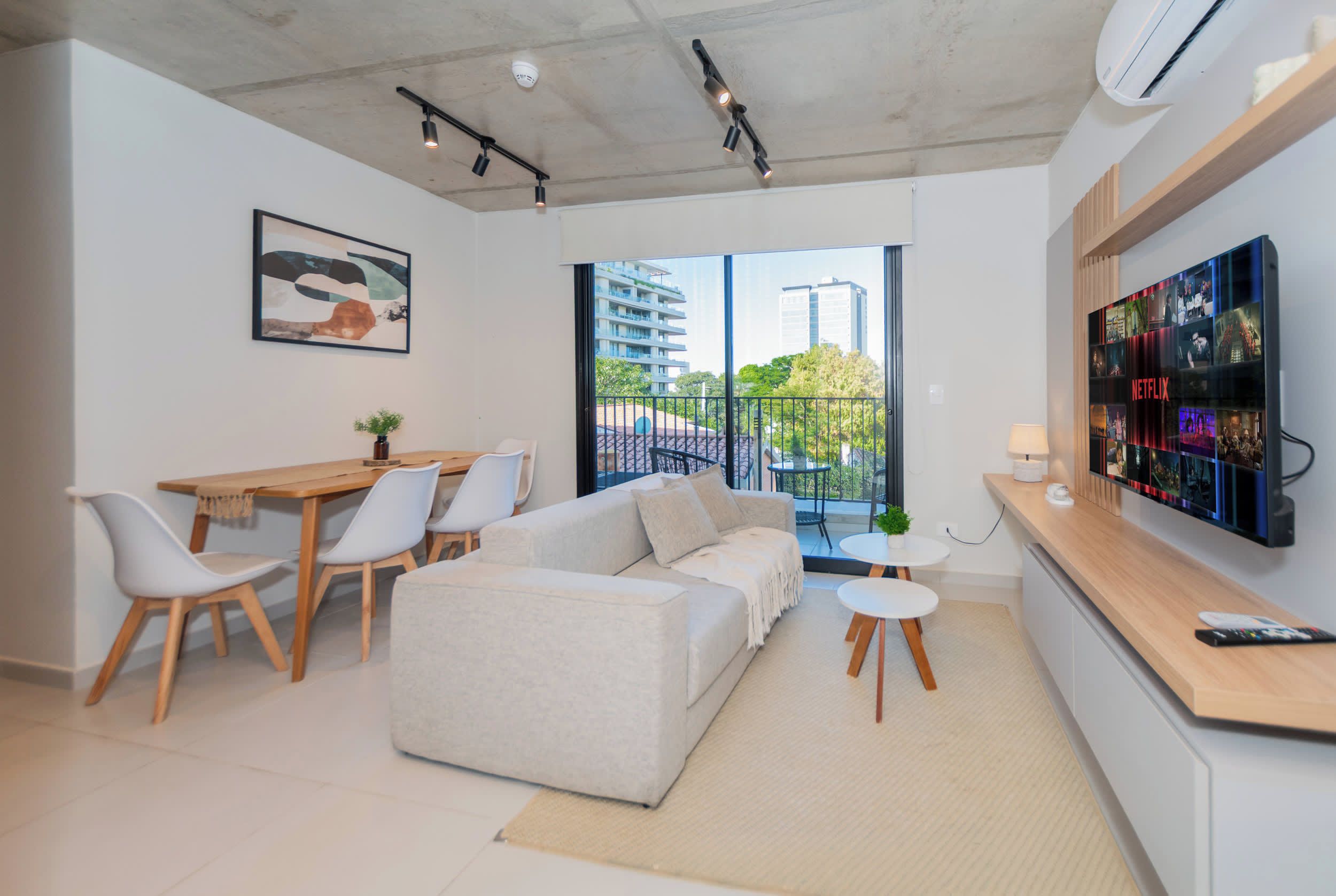 Uma sala de estar moderna com sofá, mesa de jantar, cadeiras, TV na parede e uma varanda com vista para edifícios.