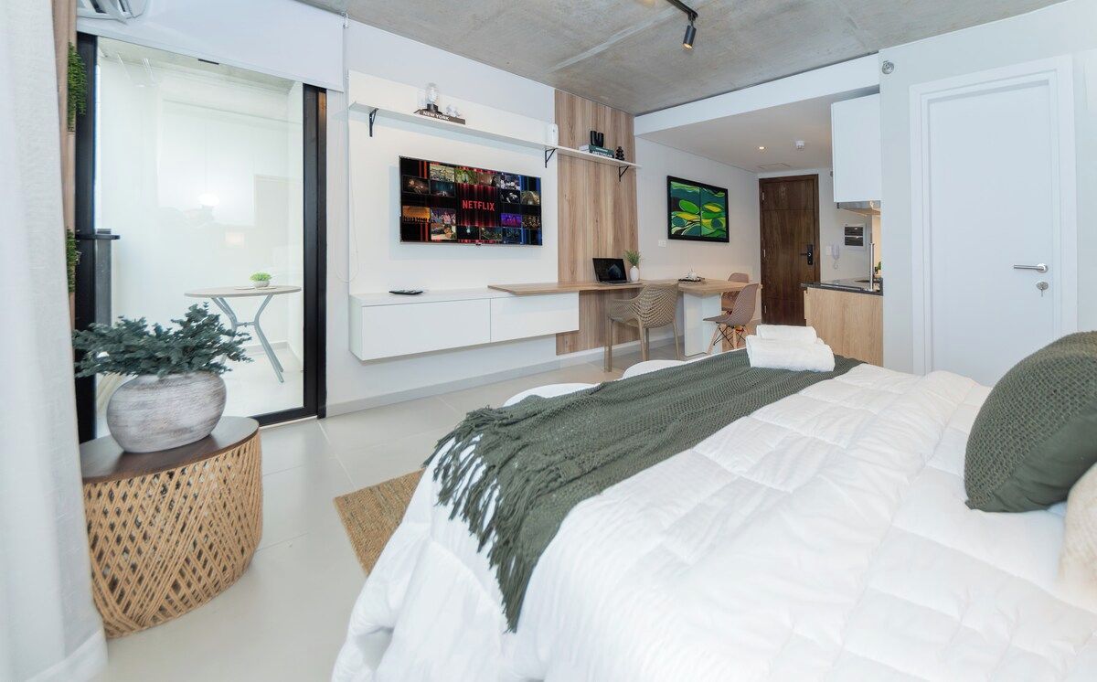 A modern bedroom with a large bed, a desk, a TV, and a sliding door leading to a balcony.