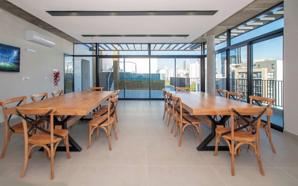A spacious meeting room with a long wooden table and chairs, large windows offering a view of the city, and a fire extinguisher on the wall.