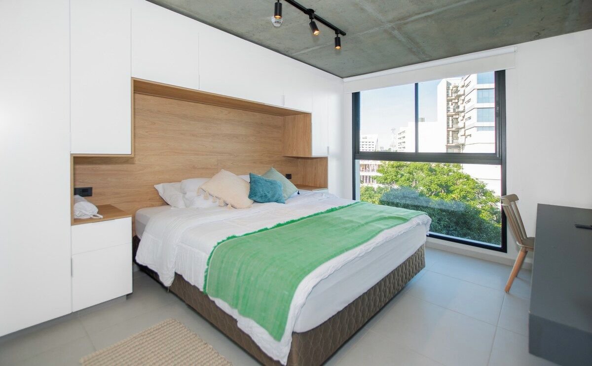 Un dormitorio moderno con cama, mesitas de noche, un escritorio y un ventanal amplio.