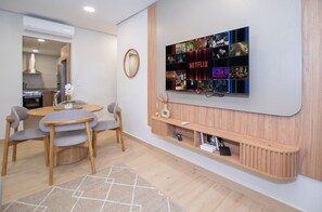 Un salon moderne avec un téléviseur à écran plat fixé au mur, un coin repas avec une table ronde et des chaises, et une cuisine visible à travers une porte ouverte.