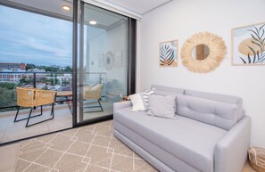 A modern living room with a grey sofa, a wicker chair, and a glass door leading to a balcony with a view of buildings. - Cozy Central Stay Near Top Malls with Pool and Gym (Asunción)