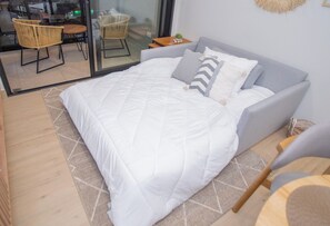 Une chambre avec un grand lit, un canapé gris et une porte en verre donnant sur un coin repas aménagé avec des meubles en osier.