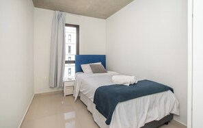 A bedroom with a bed, a nightstand, a window with a curtain, and a view of buildings outside. - Business Living, Pool & Workspace, Malls Nearby (Luque)
