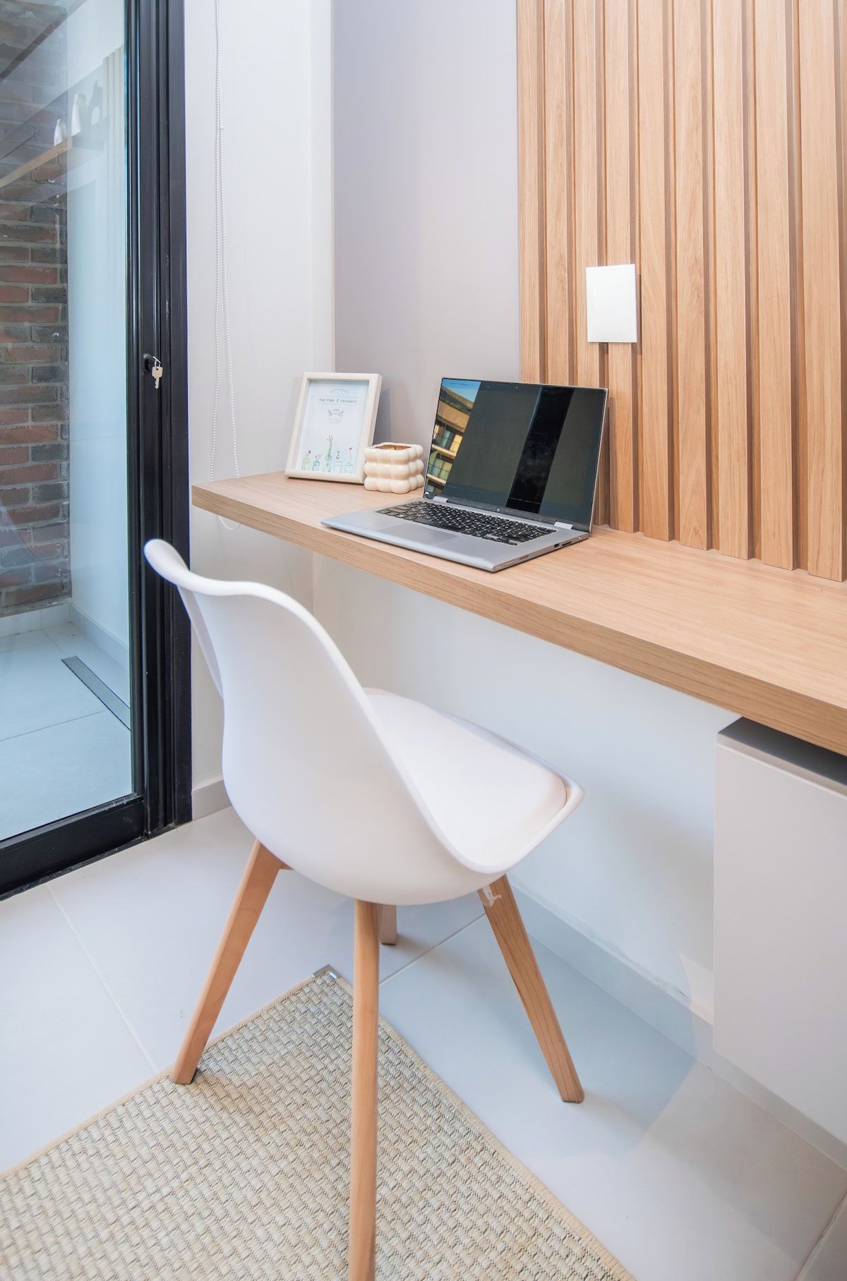 A modern workspace with a laptop on a wooden desk, a white chair, and a framed picture.