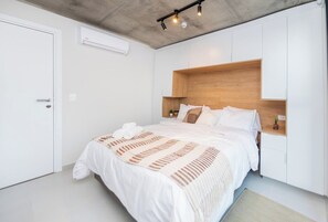Ein Schlafzimmer mit einem Bett, weißen Wänden, einem hölzernen Kopfteil und einer Klimaanlage.