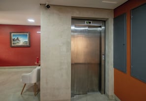 Um elevador com visor digital, uma parede vermelha com um quadro emoldurado e uma cadeira branca.