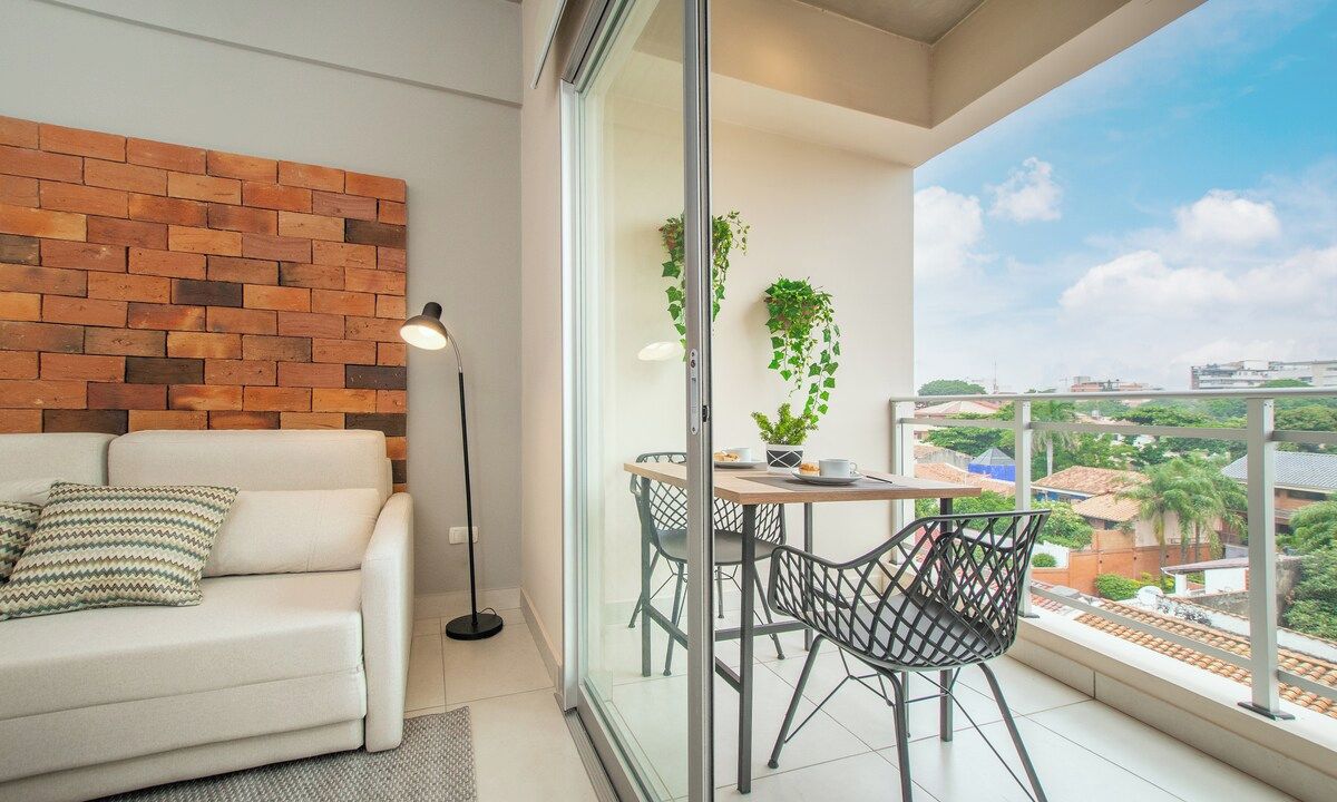 A balcony with a glass door, a white sofa, a table with chairs, and a view of buildings and greenery.