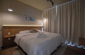 Quarto de hotel com uma cama grande, cabeceira de madeira, mesa de cabeceira e uma janela com cortinas.