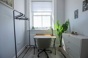 Um escritório pequeno com mesa, cadeira, radiador, janela e um vaso de planta.