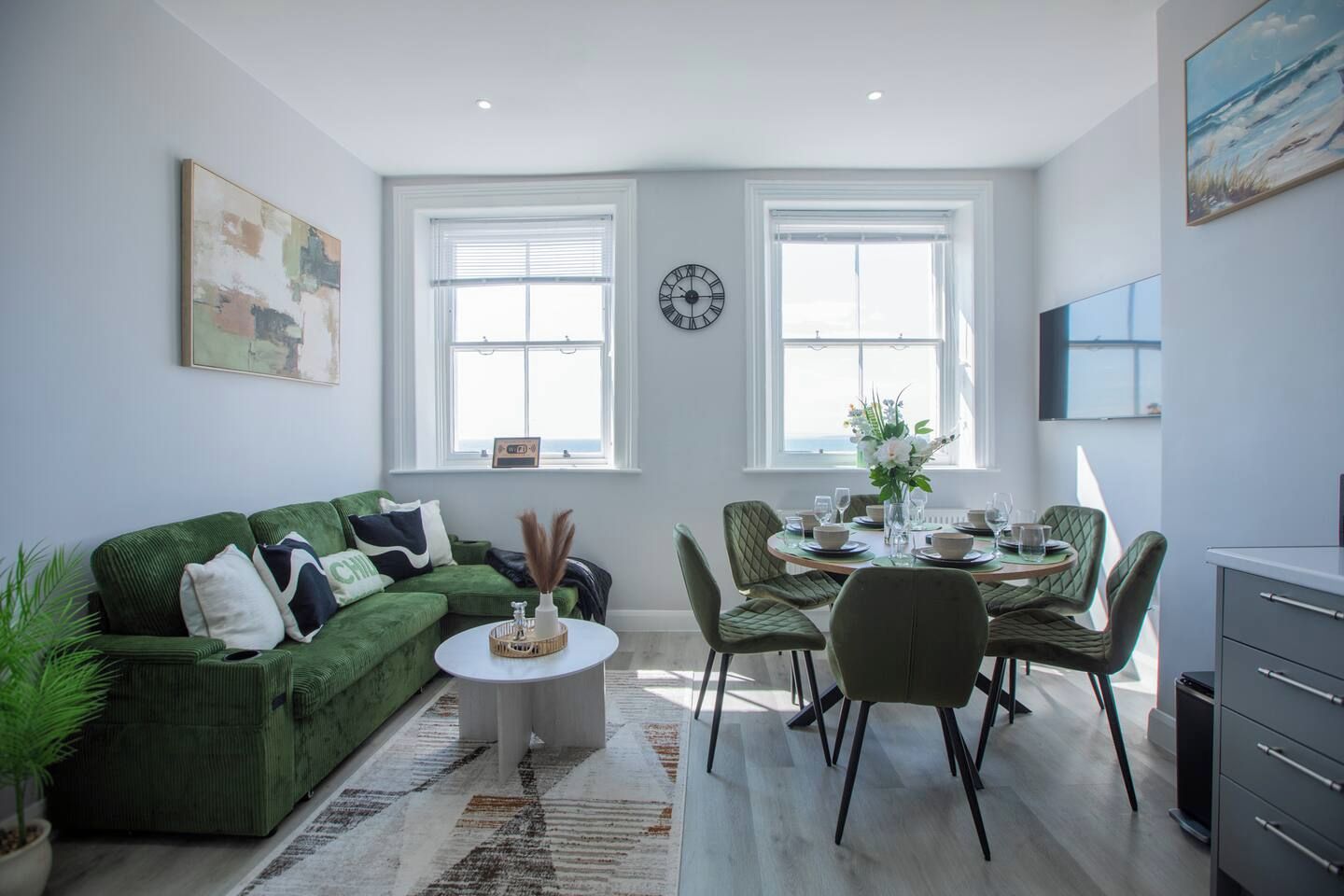 Ein modernes Wohnzimmer mit einem grünen Sofa, einem Esstisch für vier Personen und einer Küchenzeile mit Schränken.