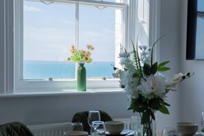 Una habitación con una mesa lista para comer, una ventana que ofrece vistas al mar y un florero con flores en el alféizar.