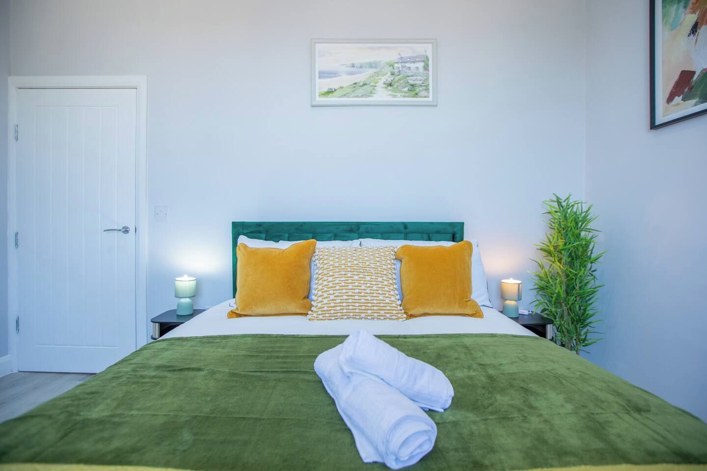 A neatly made bed with a green bedspread, two orange pillows, and a white towel. There is a framed picture on the wall above the bed, a door to the left, and a nightstand with a lamp and a plant to the right.