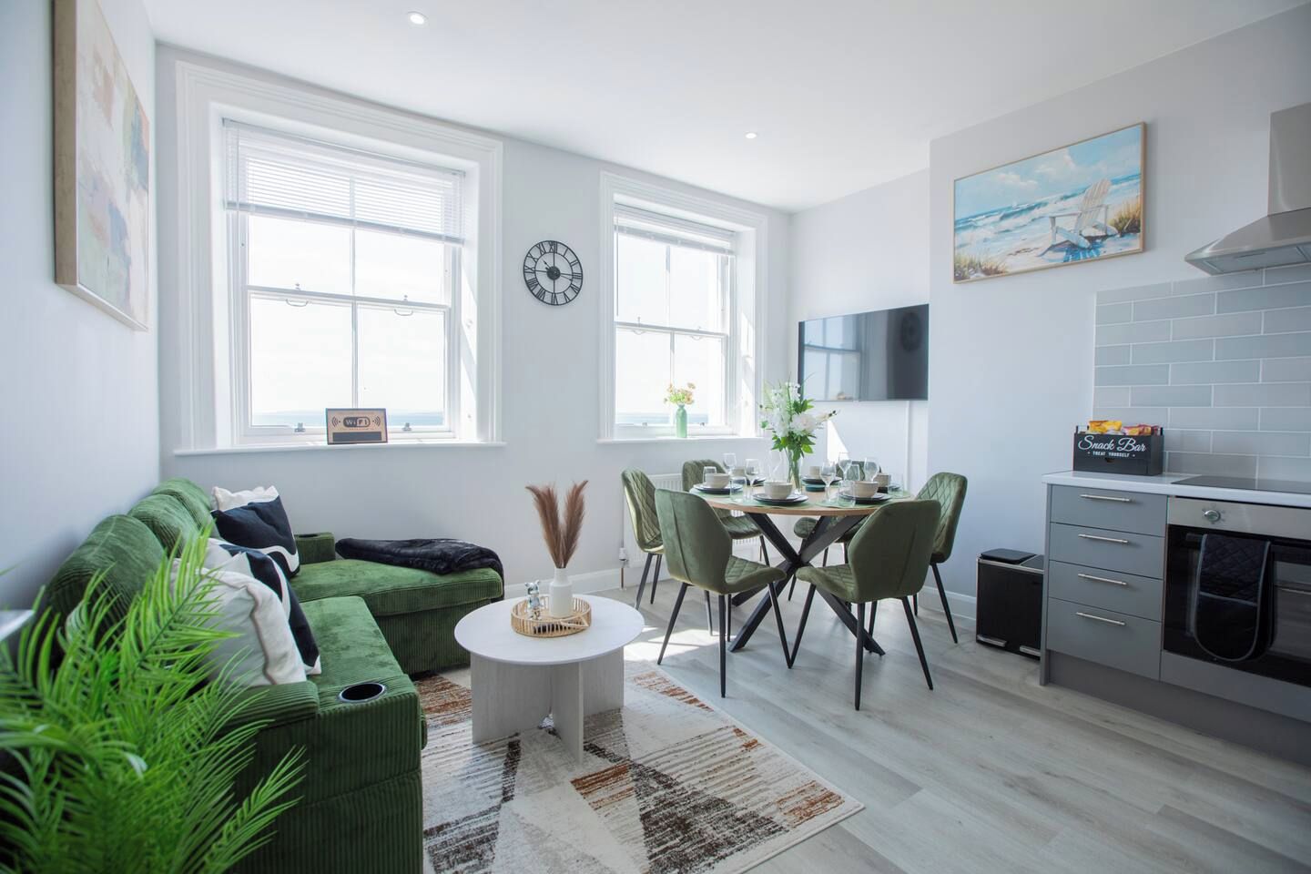 A modern living room with a green sofa, a dining table with chairs, a kitchen area with a stove, and large windows.