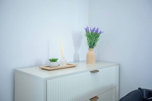 Um armário branco com uma gaveta, uma pequena planta em vaso e um vaso com flores roxas em cima.