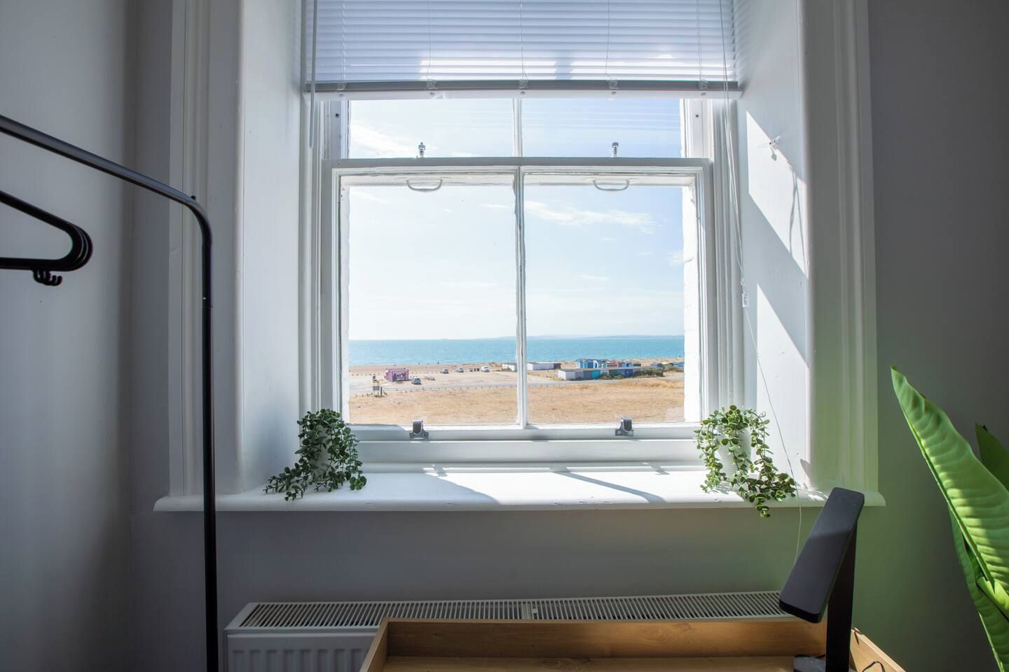 Ein Zimmer mit großem Fenster mit Blick auf einen Strand, einem Heizkörper darunter und einer Pflanze am Fensterbrett.