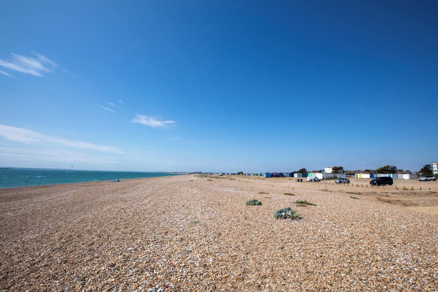 Ein Kiesstrand mit klarem Himmel, vereinzelten Gebäuden und ruhiger See.