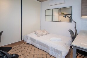 Ein kleines, ordentlich eingerichtetes Schlafzimmer mit einem Einzelbett, einem Schreibtisch, einem Stuhl und einem Spiegel an der Wand.