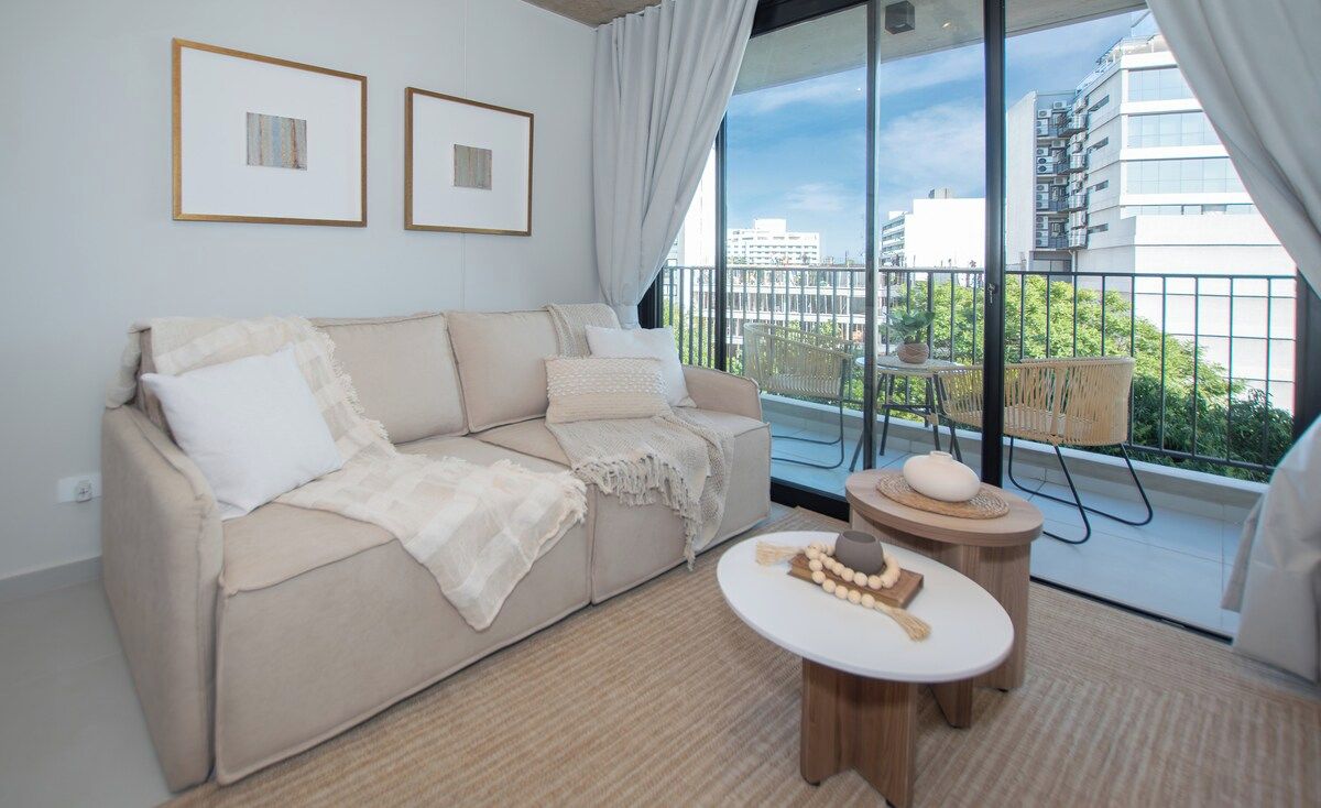 Uma sala de estar moderna com um sofá, uma mesa redonda pequena e vista para uma varanda com mobiliário de exterior.