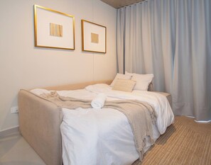 Ein ordentlich bezogenes Bett mit weißen Laken und einer beigen Decke, eine Wandsteckdose und zwei gerahmte Kunstwerke darüber.