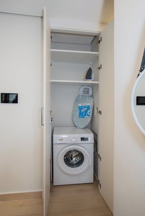 A compact laundry room with a washing machine, a drying rack, and a small shelf with a folded item. - Chic One-BR in a Prime Location with Pool and Gym (Asunción)